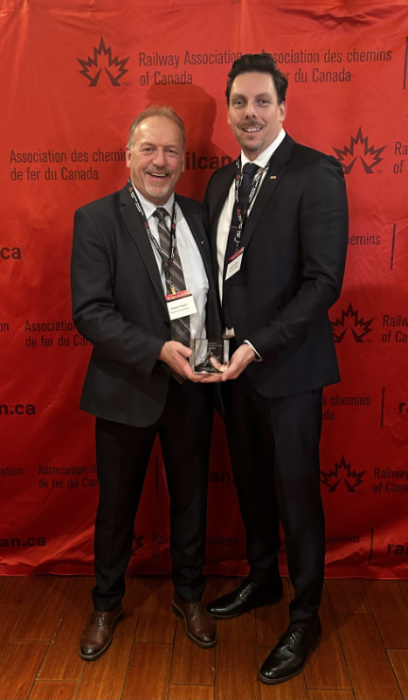 An image of two gentlemen holding a safety award in front of a red Railway Association of Canada background